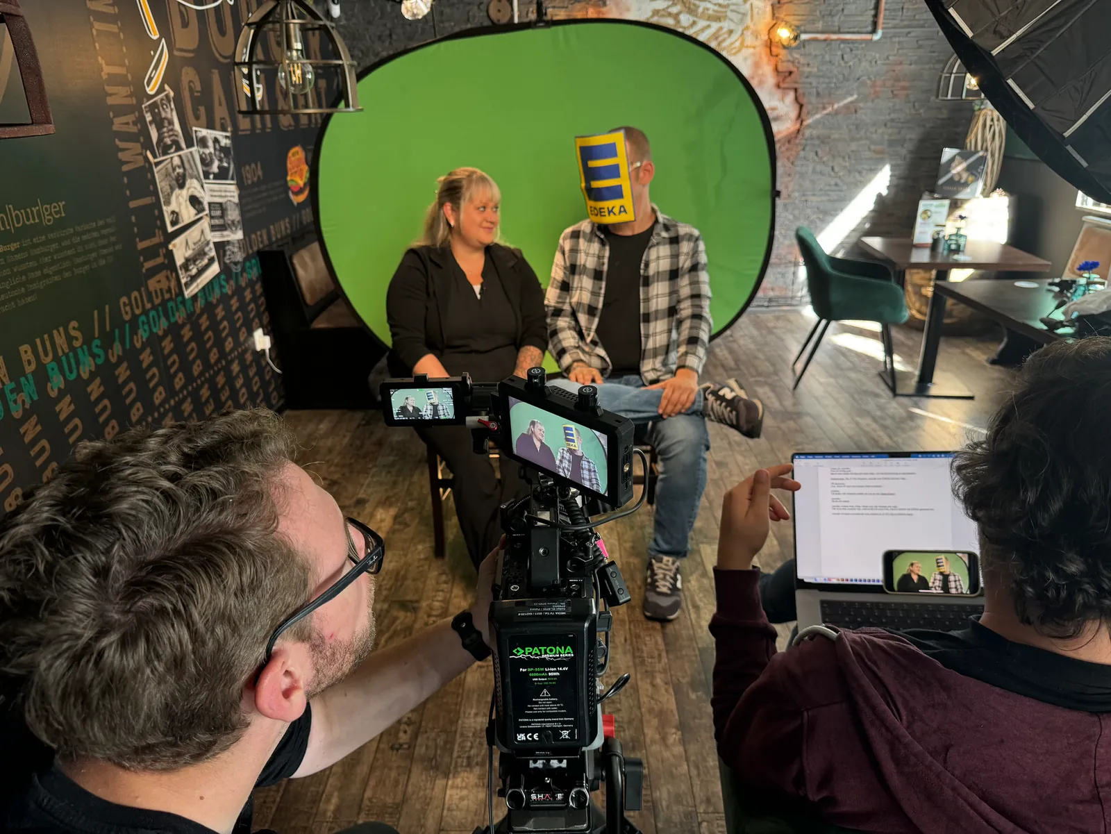 Interviewdreh vor einem mobilen Greenscreen: zwei Gesprächspartner:innen vor der grünen Wand, davor eine Kamera mit Kontrollmonitor und ein aufgeklappter Laptop