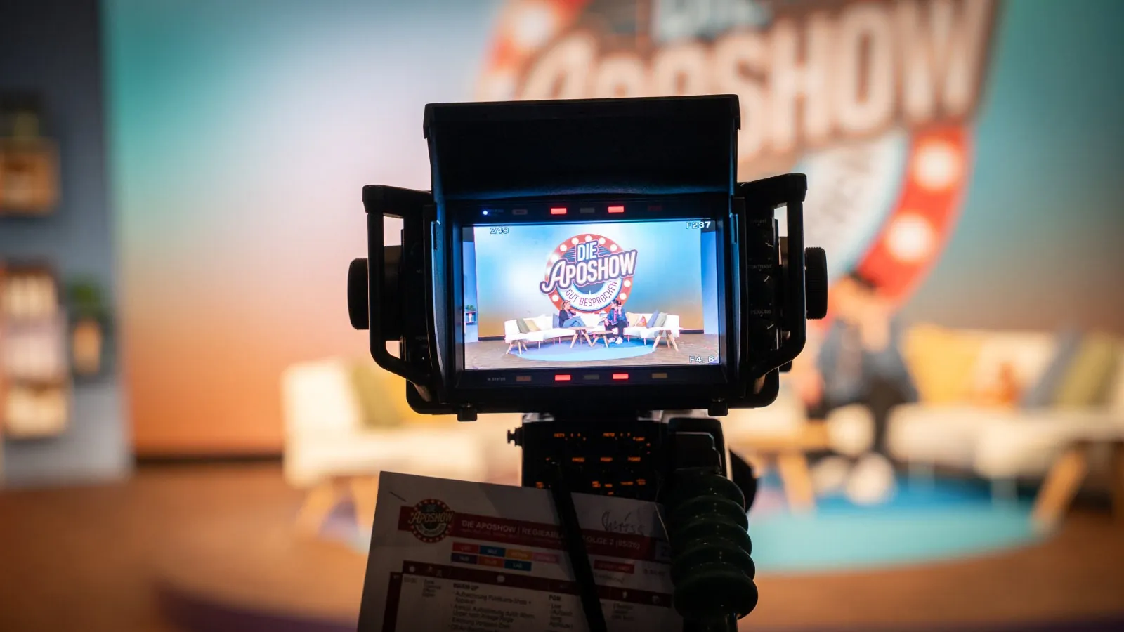 Kameramonitor mit Blick auf die ApoShow-Bühne mit Sofa und Gästen, am Stativ der Regieablauf