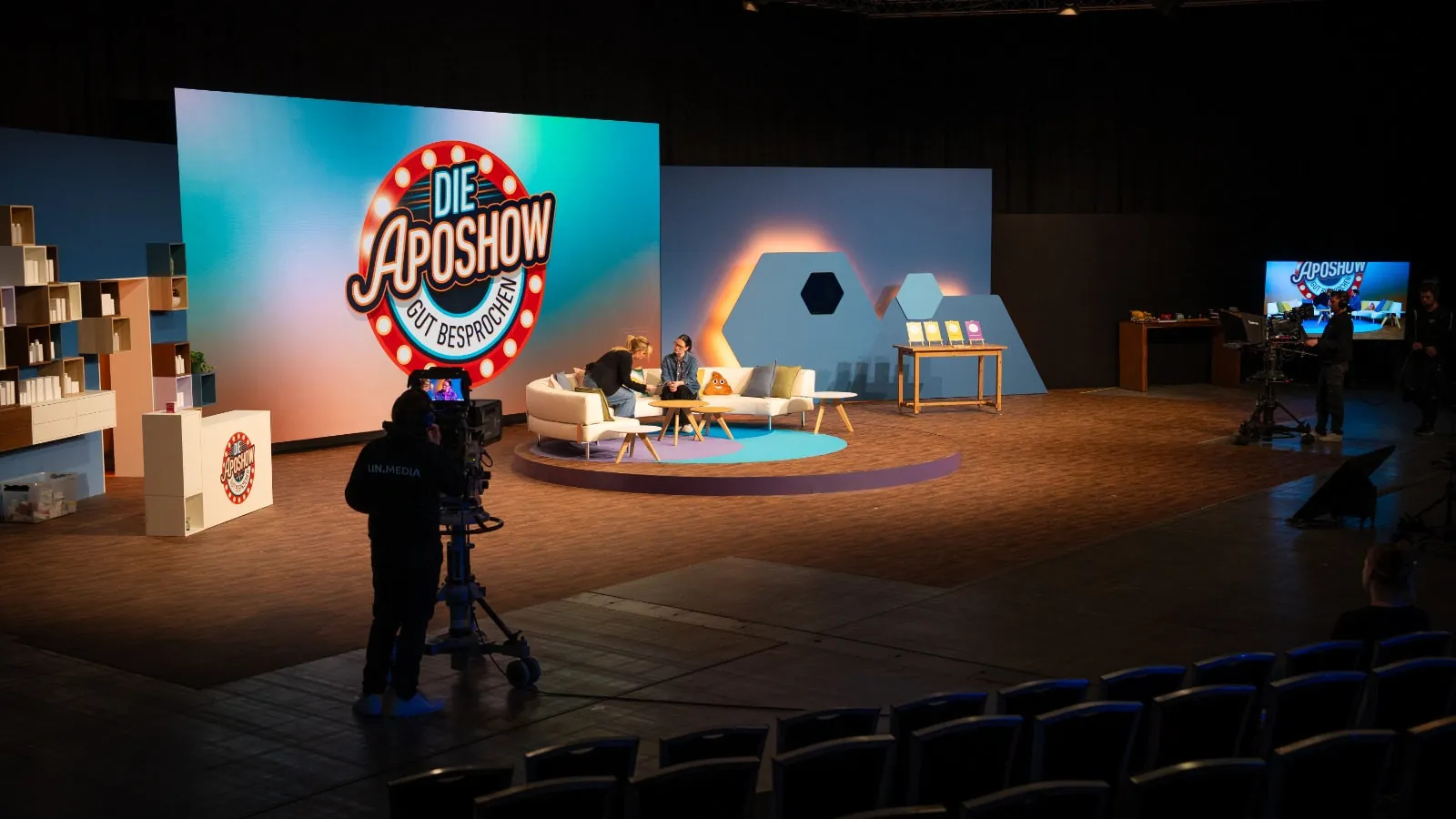 ApoShow-Bühne mit LED-Wand und Logo, Sofa für die Gäste und Studiokamera auf Pedestal, davor leere Zuschauerreihen