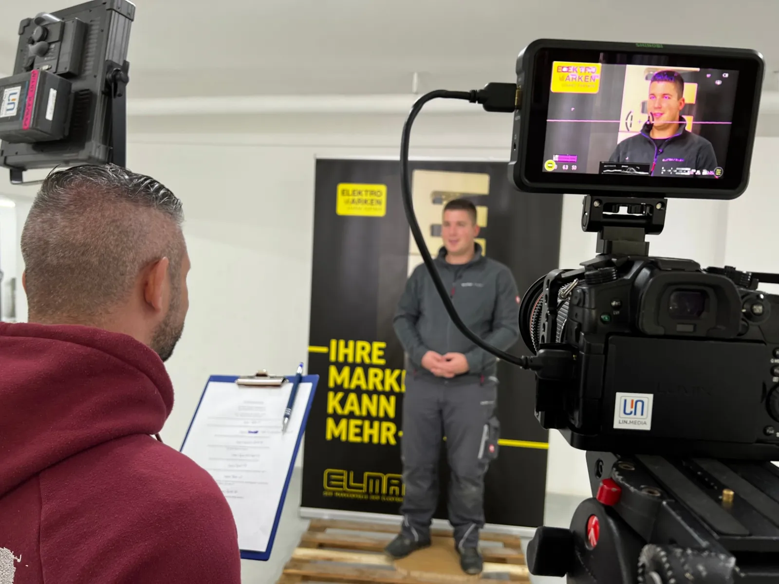 Interviewdreh im Betrieb: Protagonist vor dem Roll-up, im Vordergrund die Kamera mit Kontrollmonitor und der Interviewer mit Fragenliste