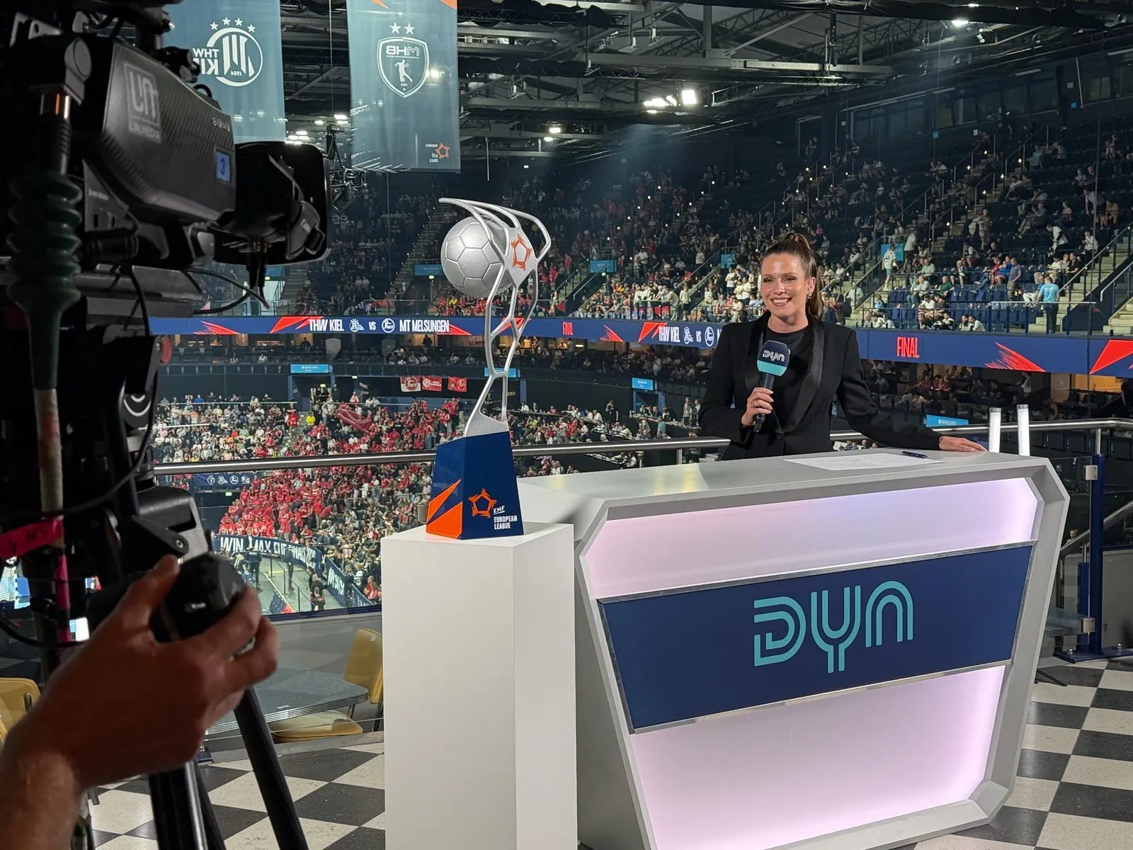 Studio-Set am Hallenrand mit Moderatorin und Pokal, davor die Kamera von LIN.Media