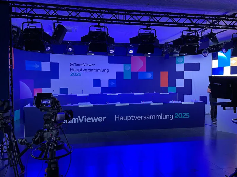 Blau ausgeleuchtete Bühne der TeamViewer-Hauptversammlung: langer Podiumstisch mit Mikrofonen vor bedruckter Rückwand, im Vordergrund eine Kamera