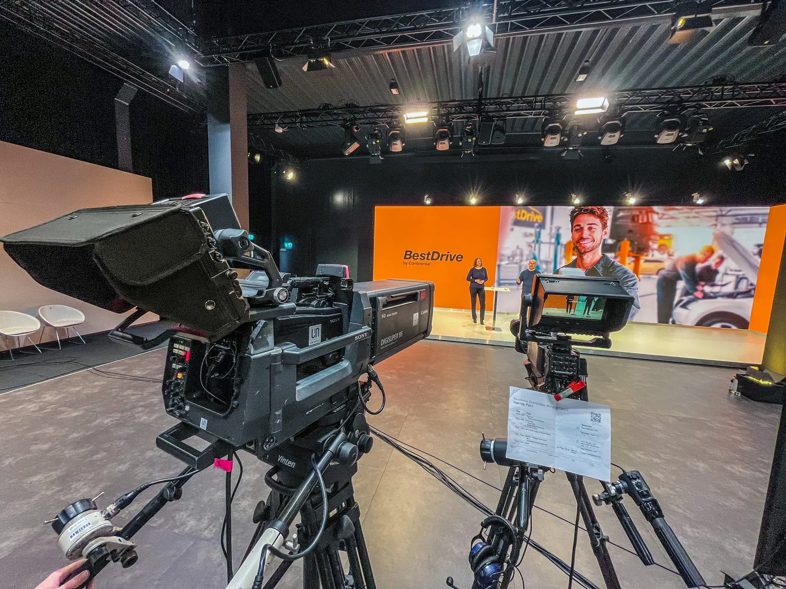 Webcast-Studio: zwei Kameras, eine davon mit Teleprompter, vor einer Bühne mit orangefarbener Rückwand, zwei Vortragende am Stehtisch
