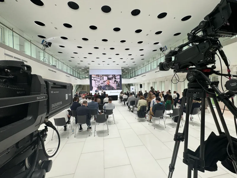 Town Hall im Atrium eines Unternehmens: Publikum auf Stuhlreihen vor einer großen Leinwand, im Vordergrund zwei Kameras am hinteren Rand des Raums